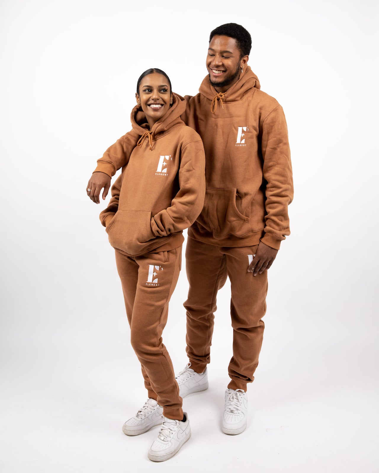 Models wearing matching light brown sweat suits. The sweat suits have the E's Element logo imprinted in white. The models are also wearing matching white sneakers. E's Element Essential Brown Sugar Sweatsuit Set by E's Element.