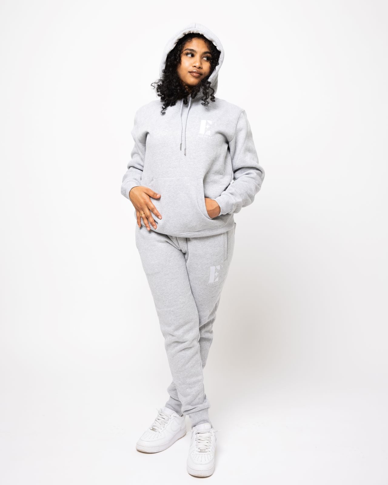Model posing and wearing a light grey sweat suit. The sweat suit has the E's Element imprinted in white.  Woman wearing grey hoodie and sweatpants.