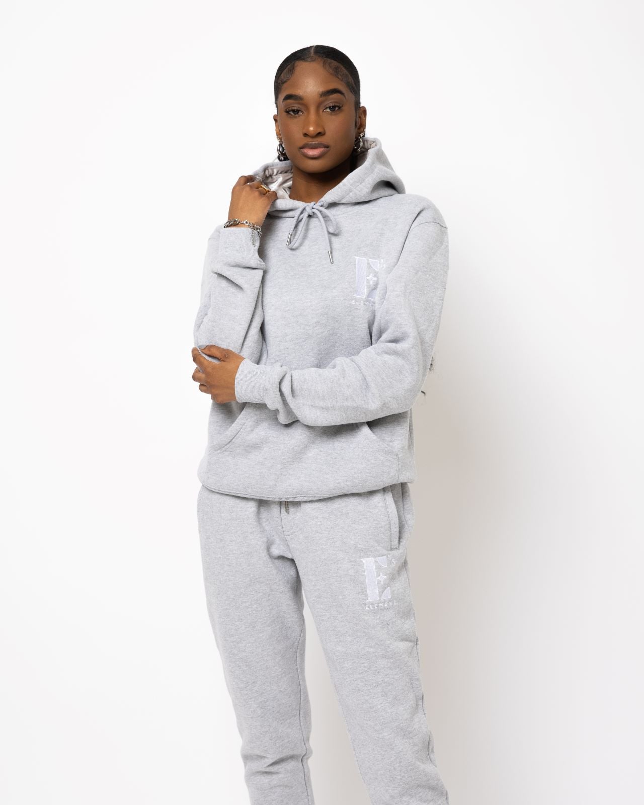 Model posing and wearing a light grey sweat suit. The sweat suit has the E's Element imprinted in white.  Woman wearing grey hoodie and sweatpants.