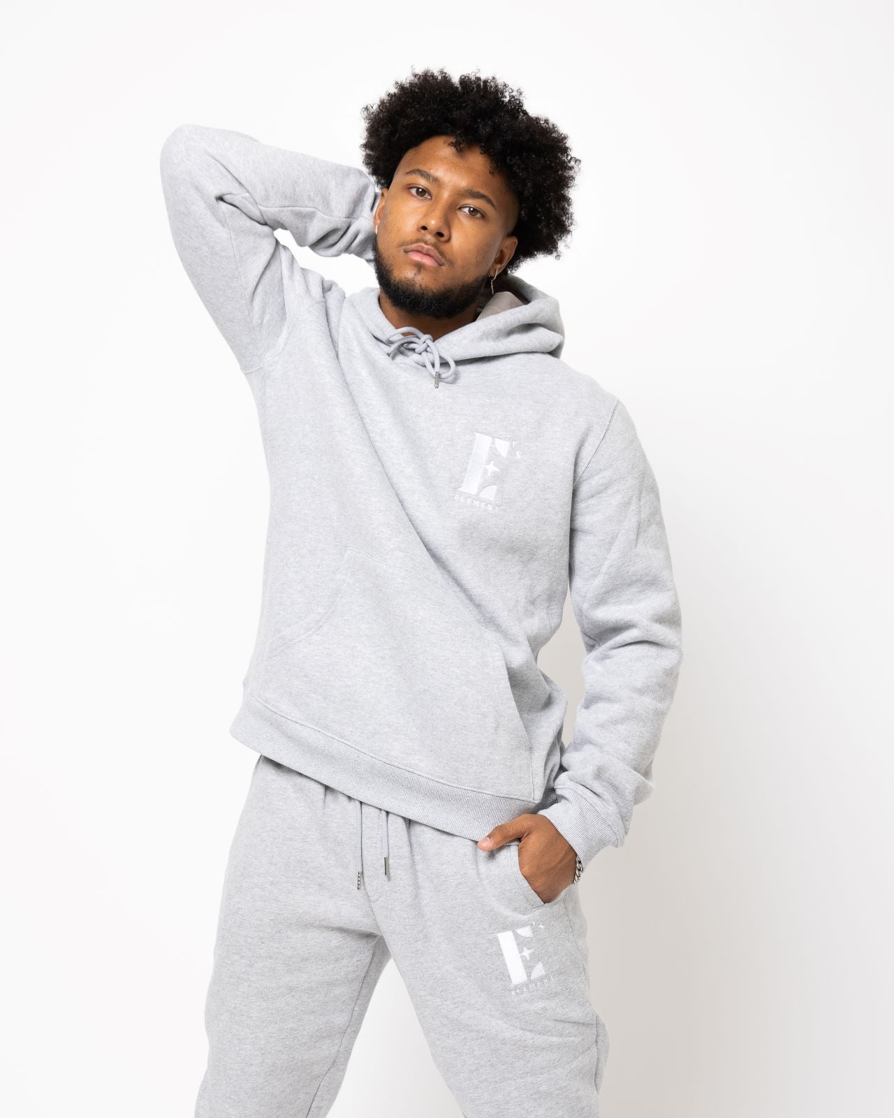 Model posing and wearing a light grey sweat suit. The sweat suit has the E's Element imprinted in white. Man wearing grey hoodie and sweatpants.