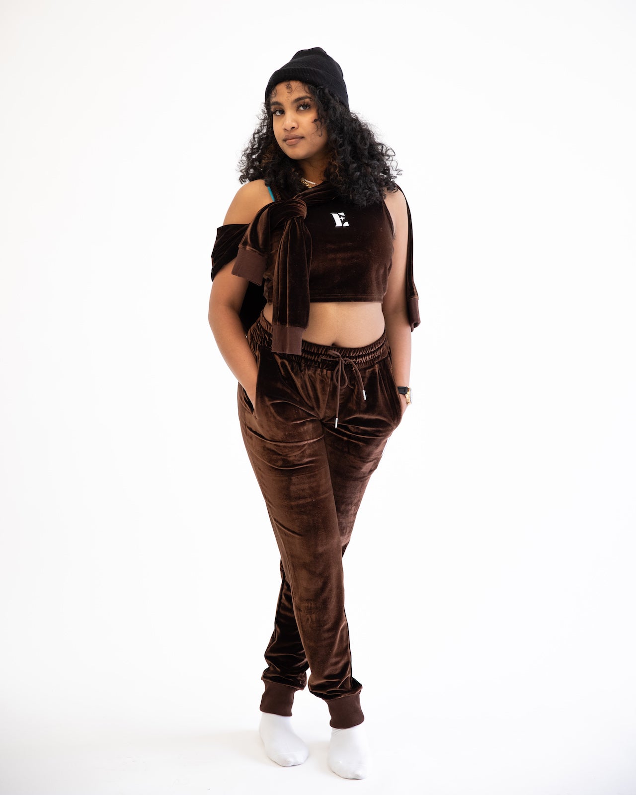 Photo of a model with curly hair posing with an e's element chocolate brown velvet crop top hoodie and jogger set. the hoodie is tied on her neck with her hands inside the pocket of the jogger set. The model is also styling her outfit with a beanie on her head and a wrist watch. Both items give the outfit an added inspired look with the e's element clothing.