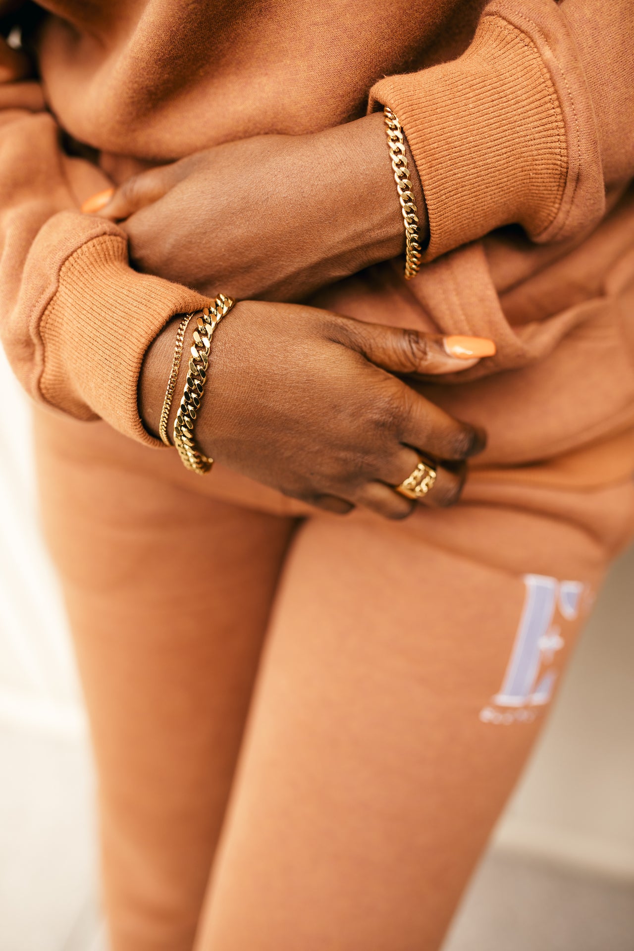 Woman wearing an orange track suit. She is wearing 18k gold stainless steel chain anklet on both wrists. Cuban Link Anklet by E's Element.