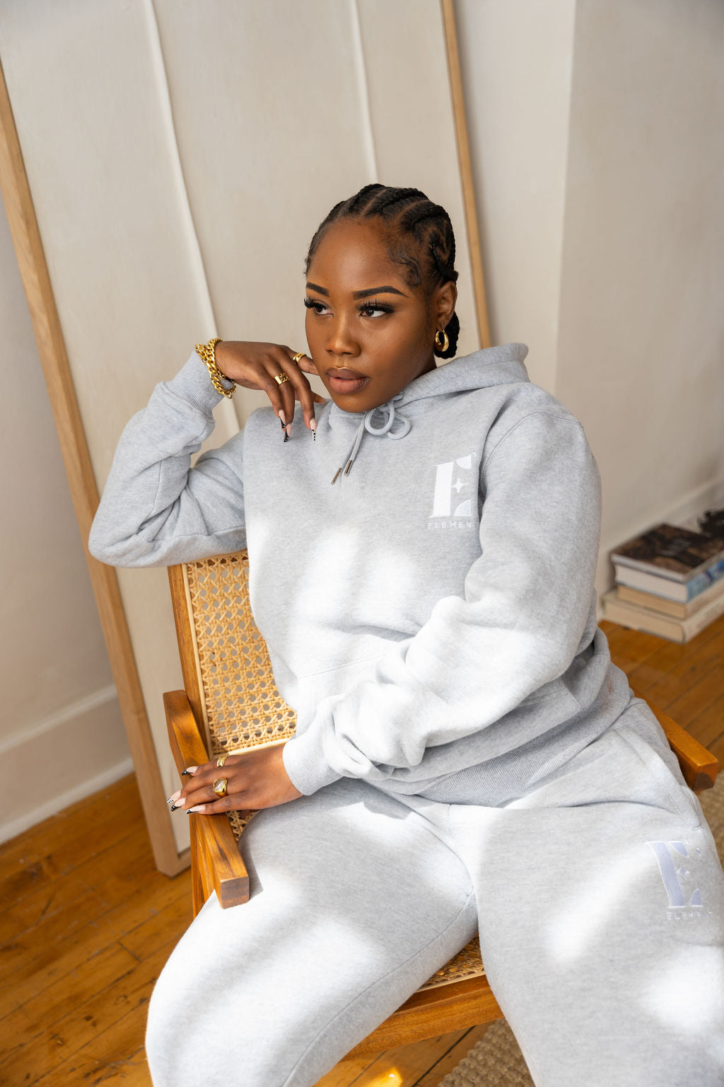 Model sitting on a chair and wearing a light grey sweat suit. The sweat suit has the E's Element imprinted in white. She is also wearing an 18k gold bracelet, rings, and earrings. Woman wearing grey hoodie and sweatpants.