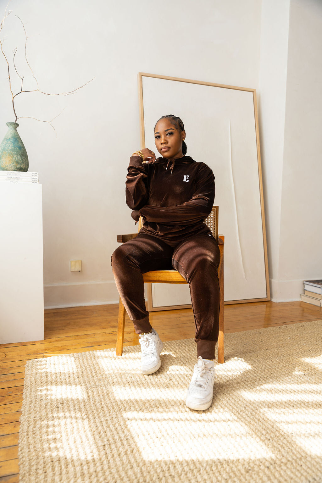 Model sitting on a wooden chair and wearing dark brown velvet hoodie and joggers. The hoodie has the E's Element logo imprinted on it. She is also wearing white sneakers. 