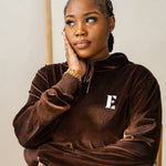 Model sitting on a wooden chair and wearing a dark brown velvet hoodie. The hoodie has the E's Element logo imprinted in white. She is also wearing 18k gold bracelets. 