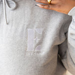 Model wearing a light grey hoodie with the E's Element logo imprinted in white. On her ring and index finger are 18k gold rings. The hoodie is a part of the Essential Light Grey Sweatsuit set by E's Element.