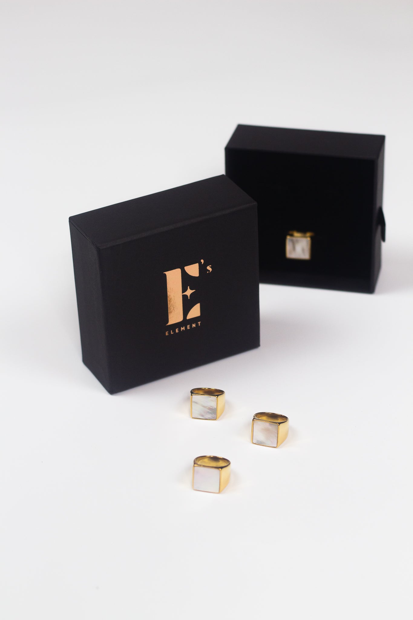 18k gold ring. The ring has a shell attached. Three of the rings are placed in front of a black container with the E's Element logo in gold. Behind the container is an open container with the ring placed inside. Shell Signet Ring by E's Element.