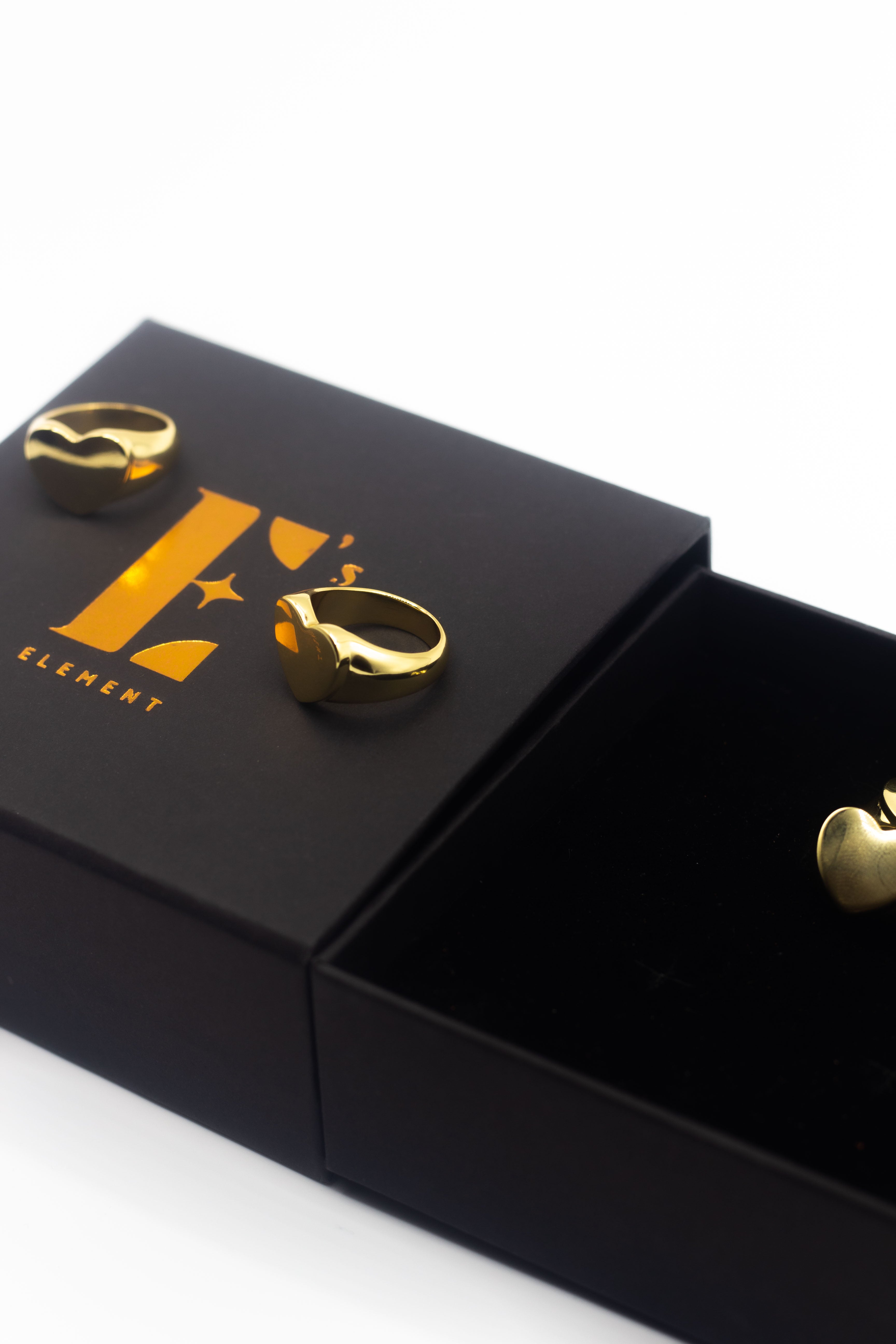 Two 18k gold heart signet rings resting on a black box with the E's Element logo imprinted in yellow. On the right is an opened black box with an 18k gold heart signet ring. Ellina Heart Signet Ring by E's Element.