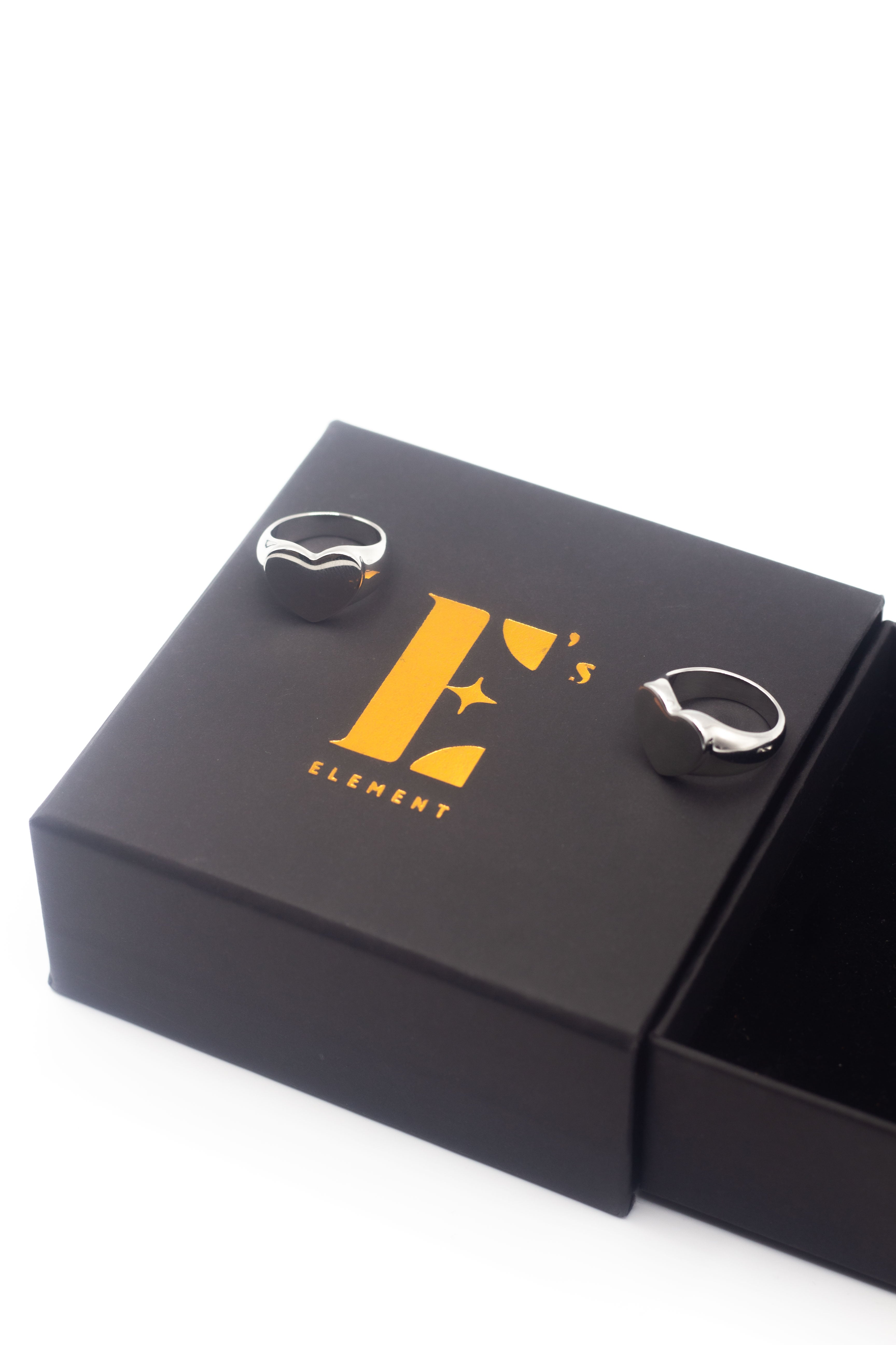 Two 18k silver heart signet rings resting on a black box with the E's Element logo imprinted in yellow. Ellina Heart Signet Ring by E's Element.
