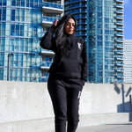 Model wearing black sweat suit. The hoodie and sweat pants has the E's Element logo imprinted on it in white. The model is also wearing white sneakers. E's Element Essential Smoky Black Sweatsuit Set by E's Element.