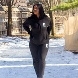 Model wearing a black sweat suit walking in the snow. The hoodie and sweat pants has the E's Element logo imprinted in white. E's Element Essential Smoky Black Sweatsuit Set by E's Element.