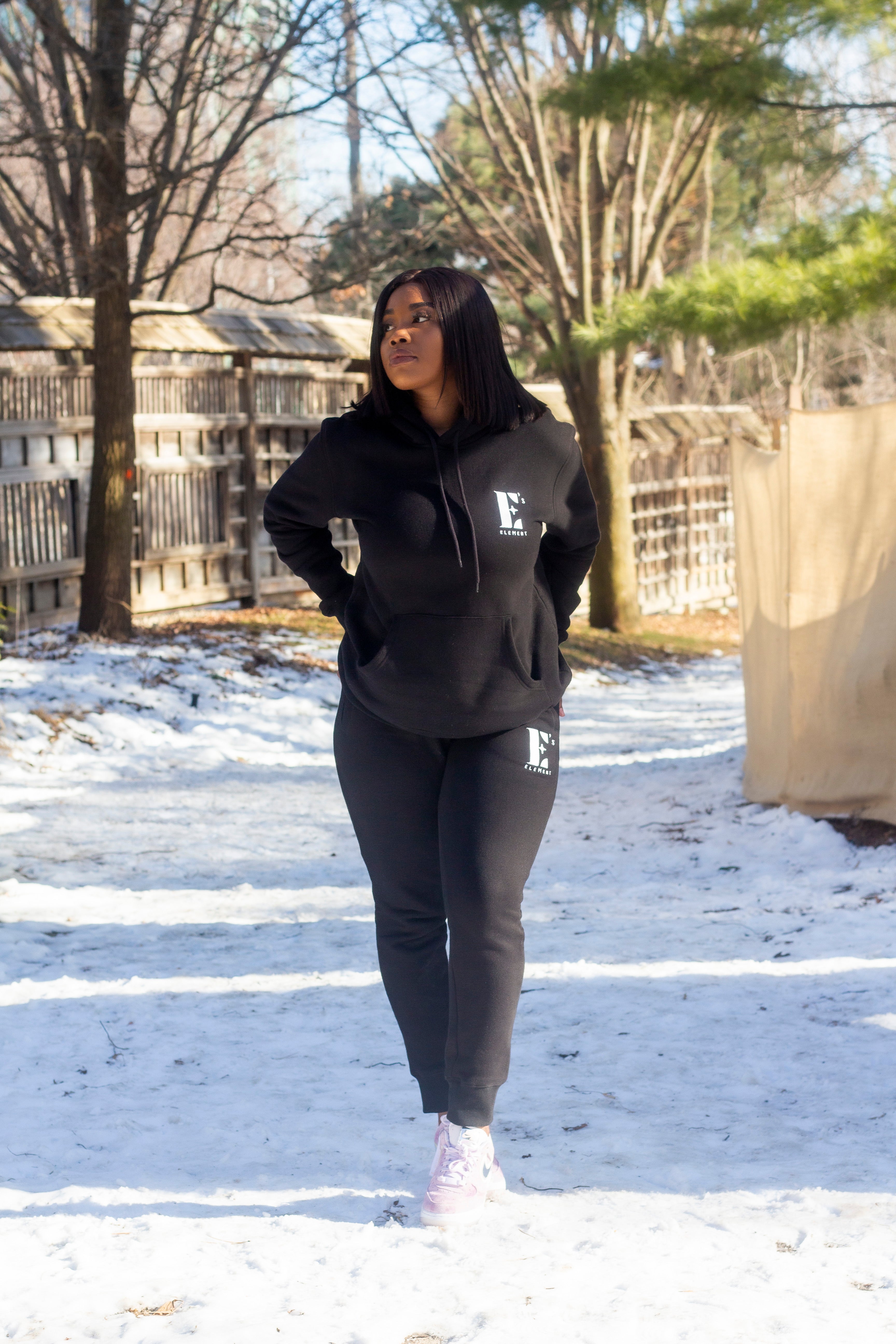Model wearing a black sweat suit walking in the snow. The hoodie and sweat pants has the E's Element logo imprinted in white. E's Element Essential Smoky Black Sweatsuit Set by E's Element.