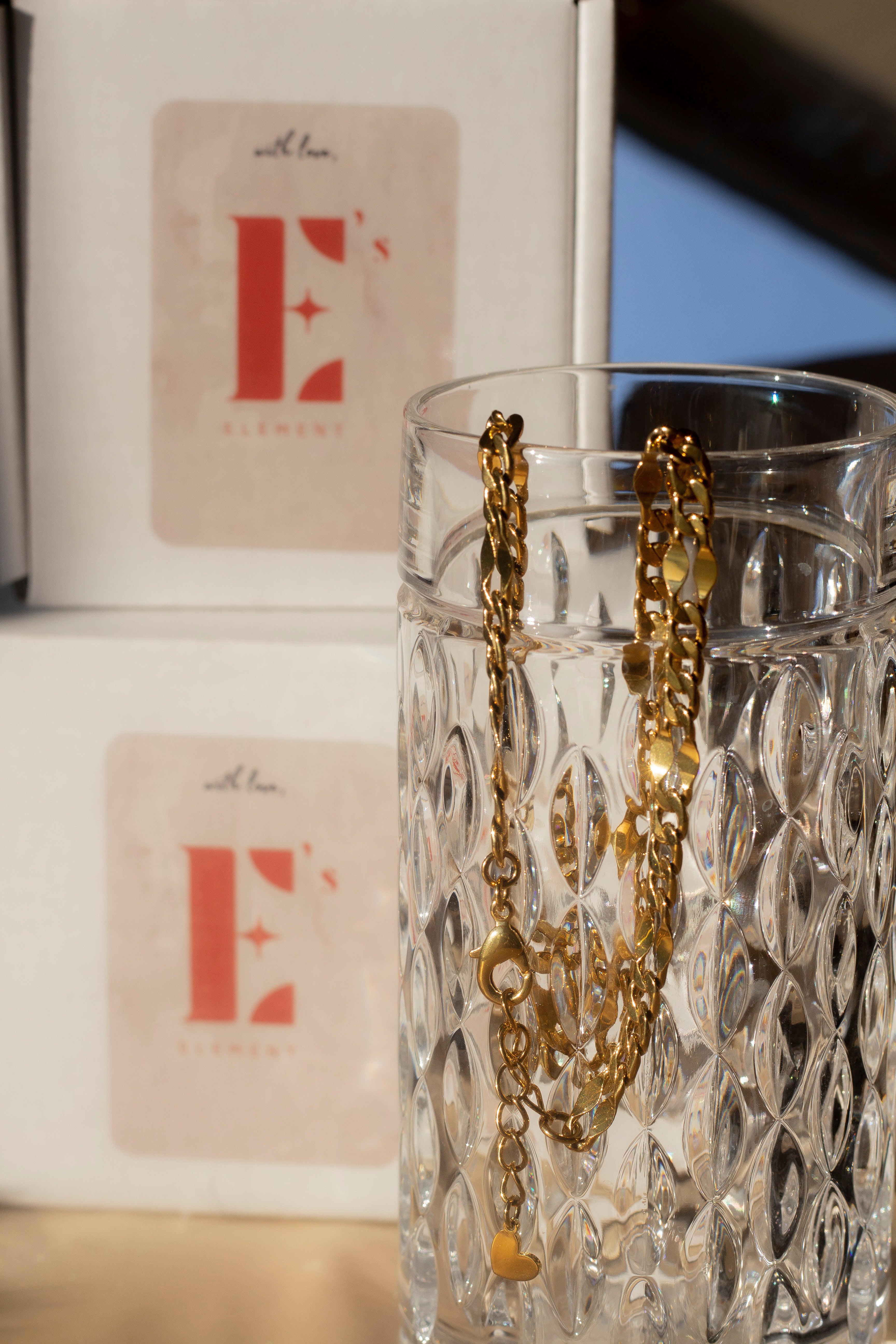 18k gold chain necklace hanging on the edge of a glass cup. Behind the cup are two white boxes stacked on each other with the E's Element logo imprinted in red in the center. Ella Figaro Necklace by E's Element.