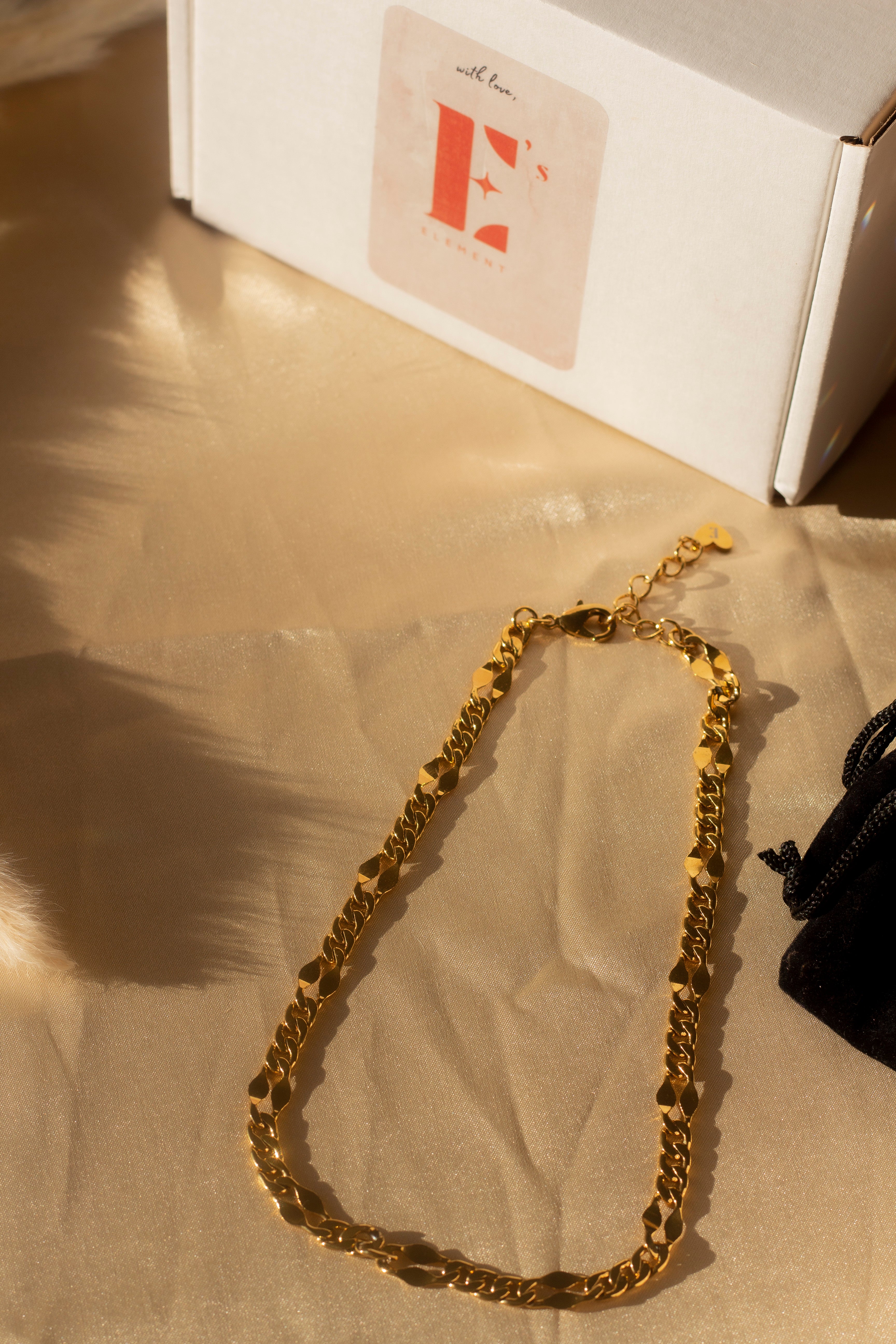 18k gold chain necklace placed on a beige cloth. Above the necklace is a white box with the E's Element imprinted in red. Ella Figaro Necklace by E's Element.