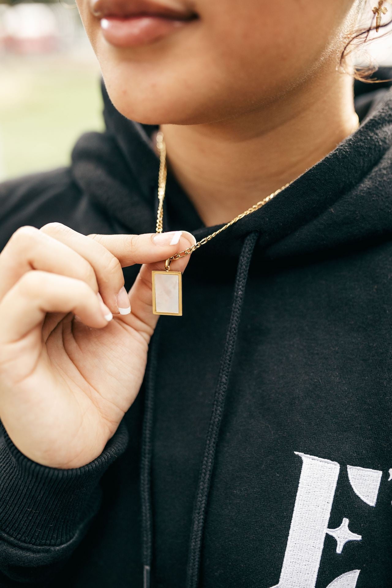 Model in a black hoodie wearing 18k gold necklace with a white rectangular charm. The hoodie has the E's Element logo in white. The Infinity Zircon Necklace by E's Element.