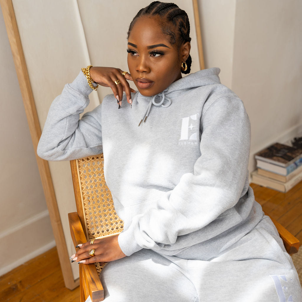 Model sitting on a chair wearing a light grey sweat suit. She is also wearing white sneakers. The sweat suit has the E's Element imprinted in white. The model is also wearing white sneakers. Woman wearing grey hoodie and sweatpants.