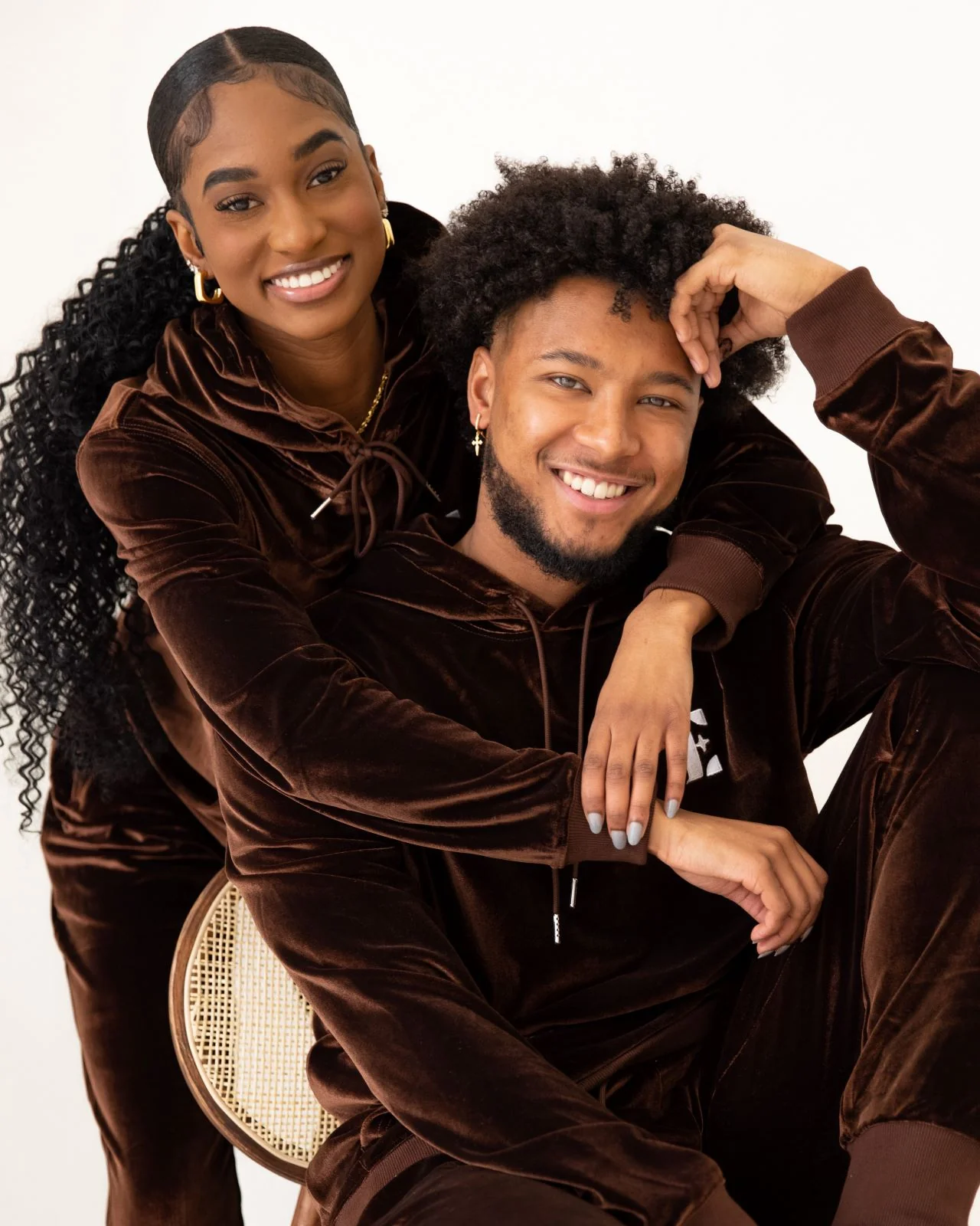 Man sitting in a chair and woman wraps her arms around him, wearing E's Element Chocolate Brown Velvet Jogger Set on white background