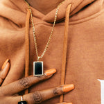 Model in an orange hoodie wearing an 18k gold ring and chain necklace. The ring has zircon stone in the middle of a rectangular black surface. The necklace has black rectangular charm with gold edges. Infinity Zircon Ring by E's Element. Infinity Zircon Ring by E's Element.