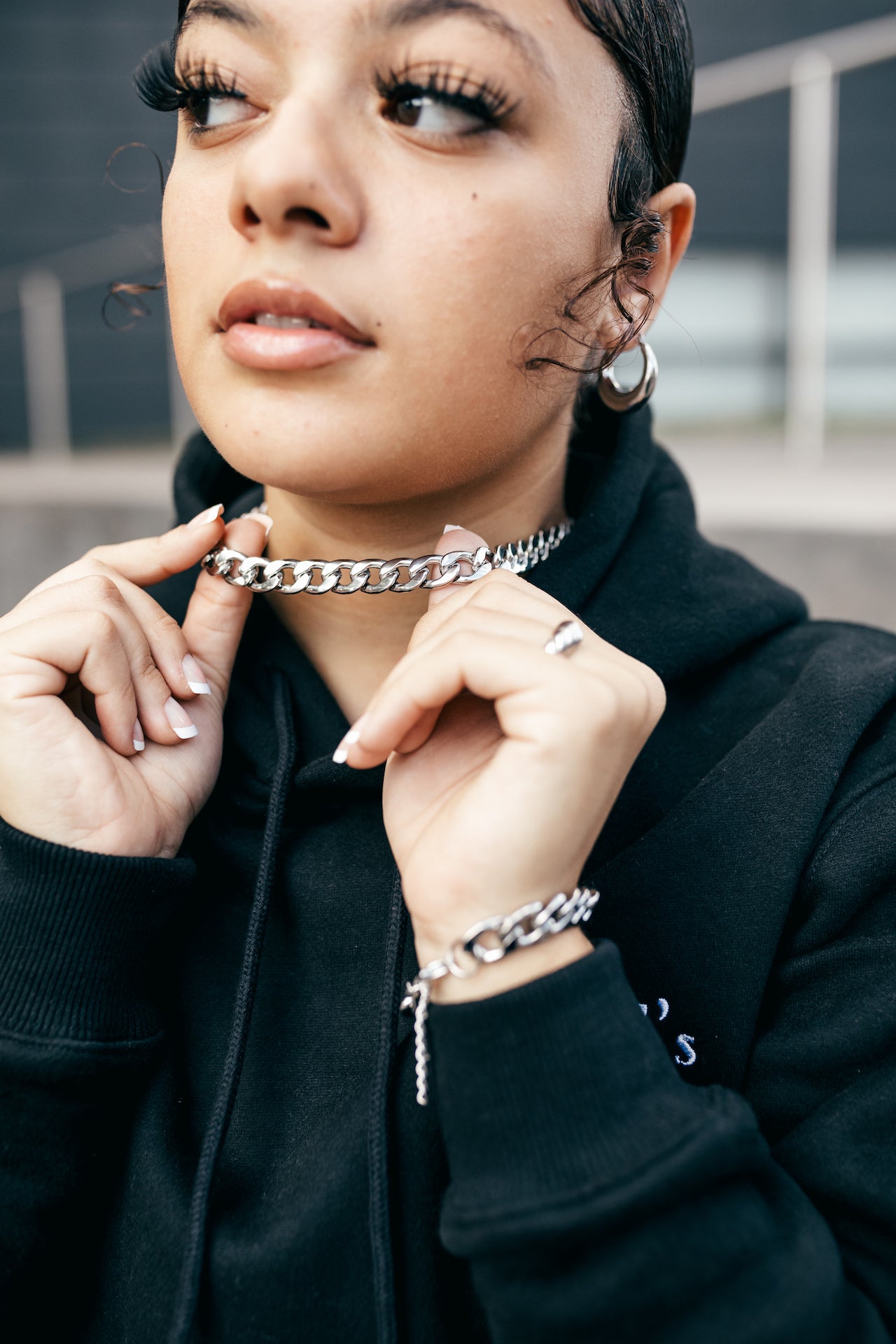 Model in a black hoodie wearing stainless steel chain necklace and bracelet. She is also wearing stainless steel rings and earrings. The Emmanuela Set in Steel by E's Element.