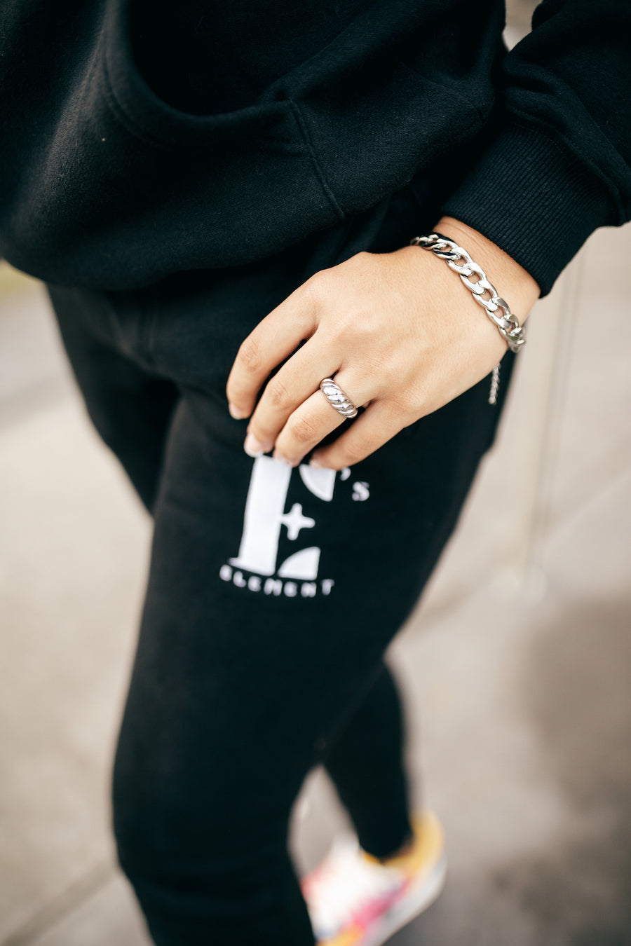 Model in a black sweat suit wearing stainless steel chain bracelet and ring. The sweat pants has the E's Element logo in white. The Emmanuela Set in Steel by E's Element.