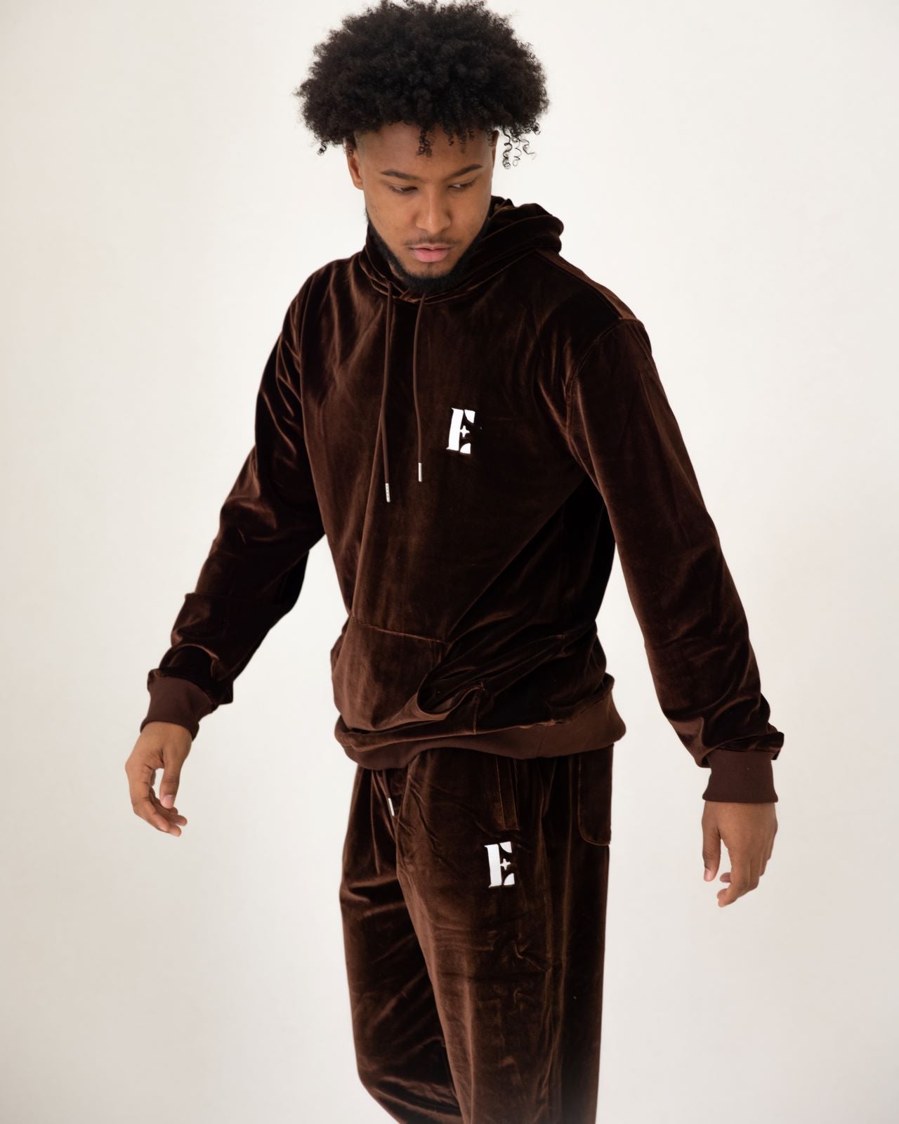 Photo of  a male model showing a closer look of the e's element chocolate brown velvet jogger set. the picture gives a closer view of the logo on the jogger set and shows the glow on the velour material used to make the set.