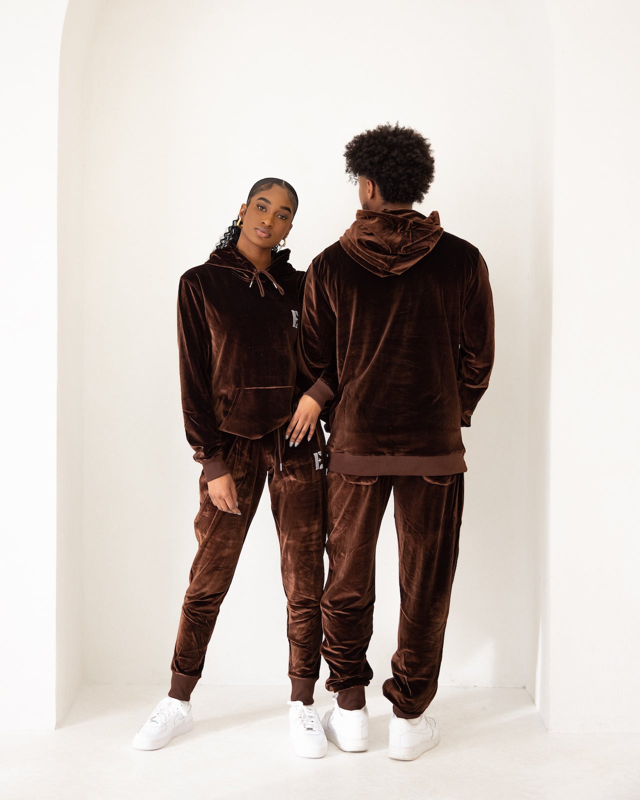 Photo of a male and female model posing in the e's element chocolate brown velvet jogger set. the female model is is showing the front view of the jogger set while the male model is showing the back view of the jogger set.