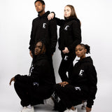 Models wearing black sweatsuits. The sweatsuits are imprinted with the E's Element logo in white. The models are also wearing matching white sneakers.E's Element Essential Smoky Black Sweatsuit Set by E's Element.