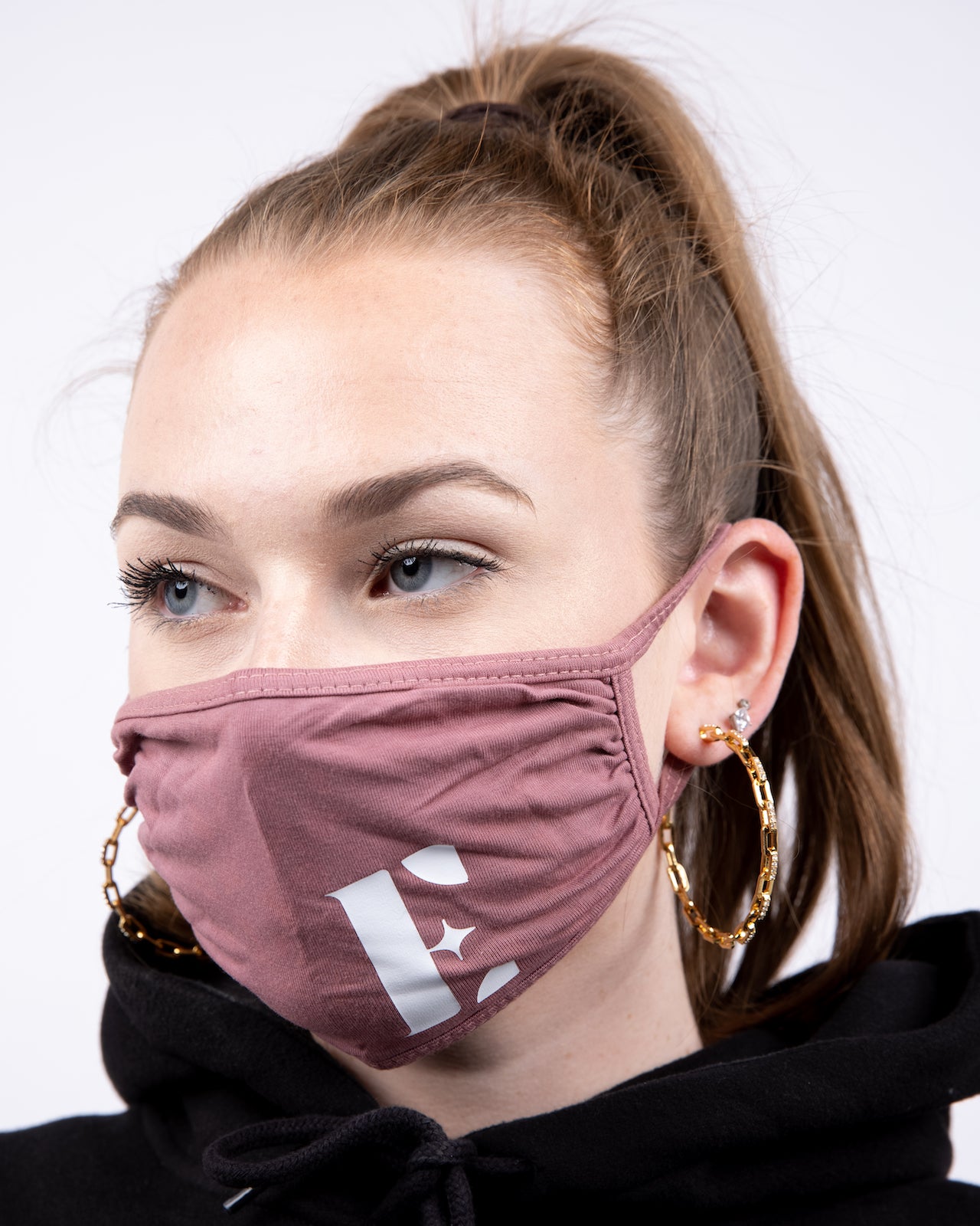 Model in a black hoodie wearing a pink face mask with the E's Element logo in white. She is also wearing gold chain hoop earrings and silver studs. Mauve Pink Face Mask by E's Element.