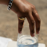 Model holding a glass of water. She is wearing an 18k gold croissant shaped ring. She is also wearing a silver chain bracelet. Thick Croissant Rings by E's Element.