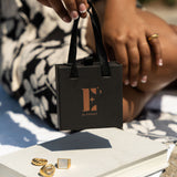 Model in a black and white dress holding a black gift box with a handle. The gift box has the E's Element logo imprinted in gold. Underneath the box are three 18k gold rings resting on a white book. Gift Box With Handle by E's Element.