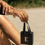 Model sitting at the beach holding a black gift box at the handle. The gift box has the E's Element logo imprinted in gold. The model is also wearing 18k gold rings, bracelet, and anklets. Gift Box With Handle by E's Element.