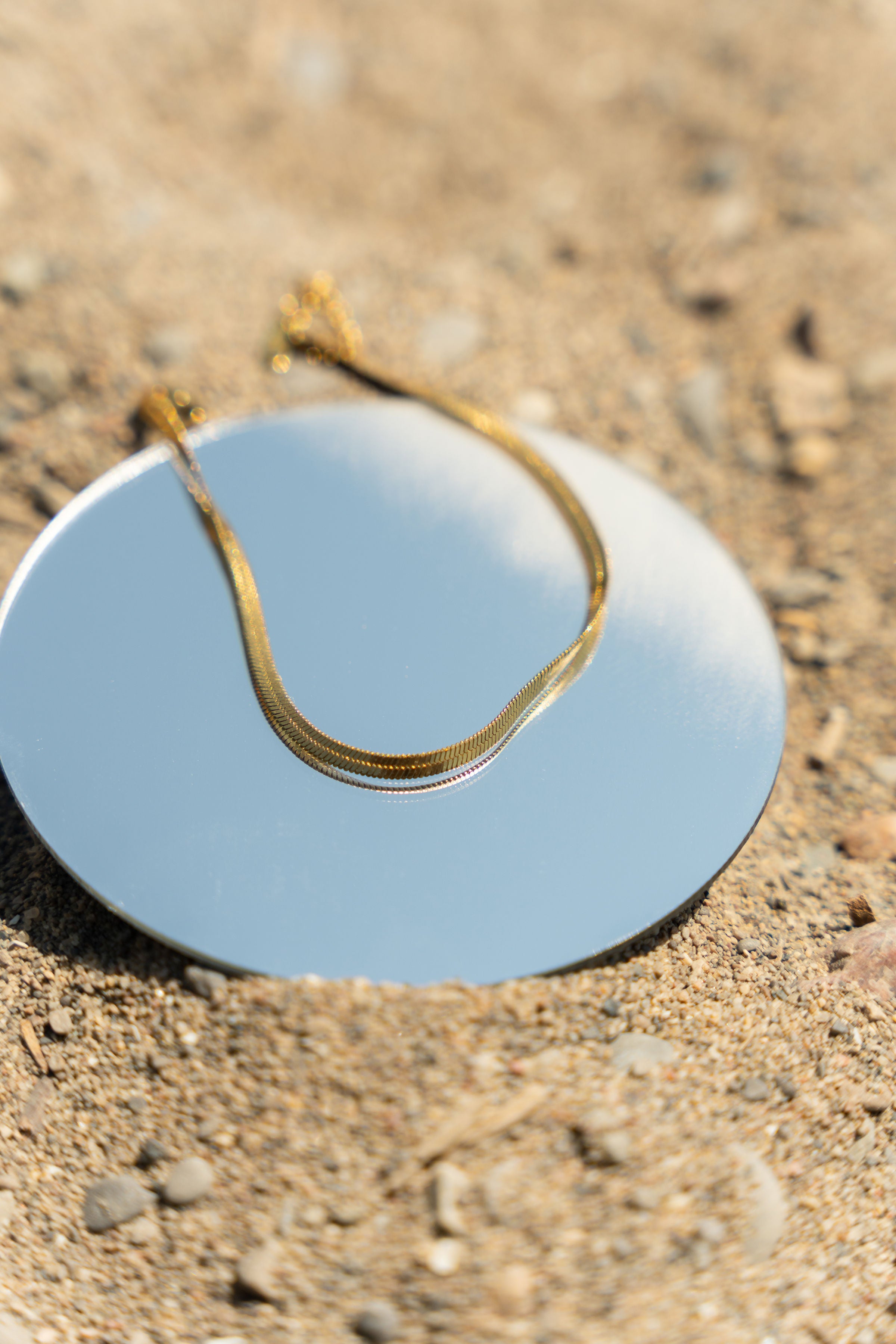 18k gold stainless steel anklet resting on top of a round mirror on the sand.