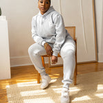 Model sitting on a chair wearing a light grey sweat suit. She is also wearing white sneakers. The sweat suit has the E's Element imprinted in white. The model is also wearing white sneakers. Woman wearing grey hoodie and sweatpants.