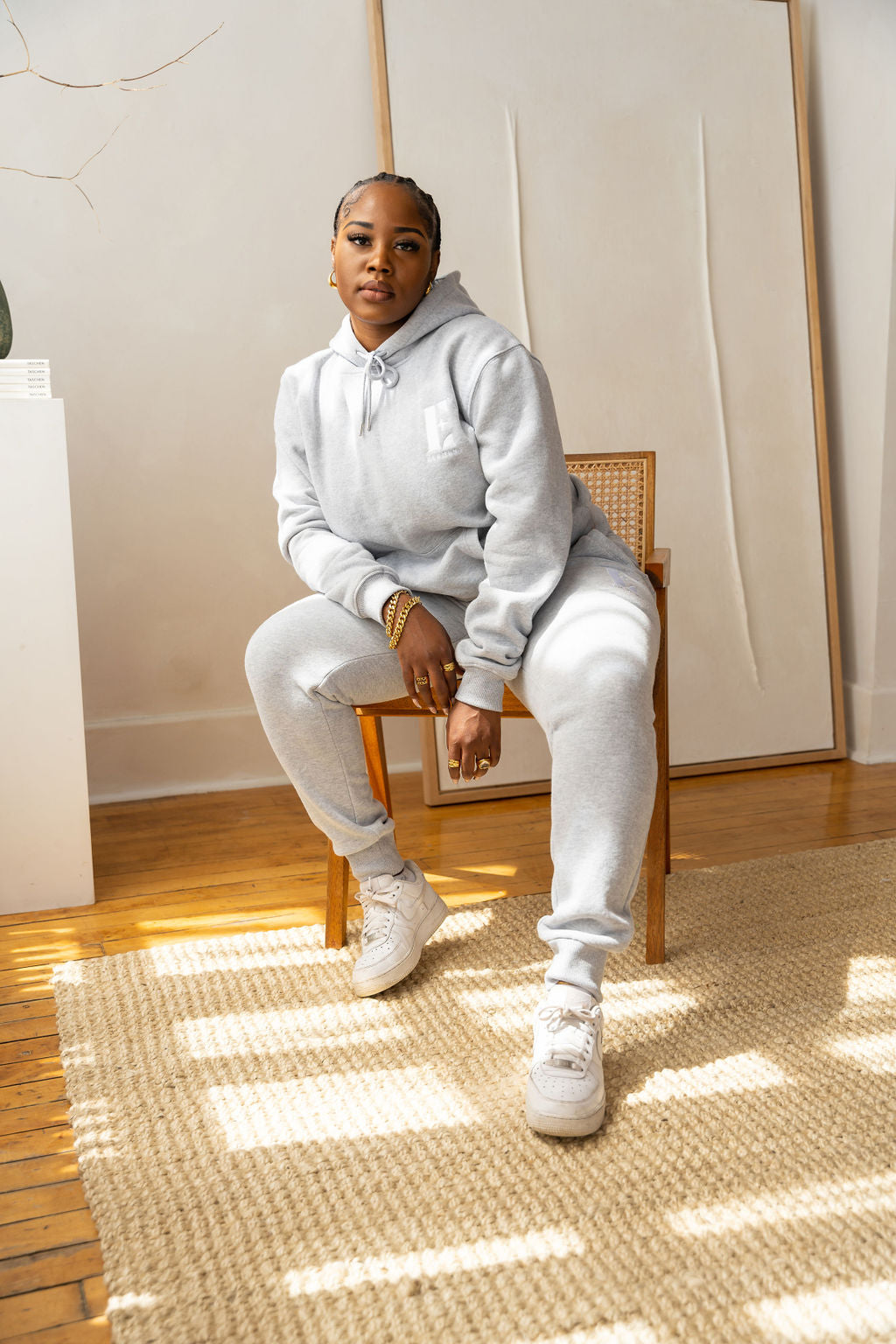 Model sitting on a chair wearing a light grey sweat suit. She is also wearing white sneakers. The sweat suit has the E's Element imprinted in white. The model is also wearing white sneakers. Woman wearing grey hoodie and sweatpants.