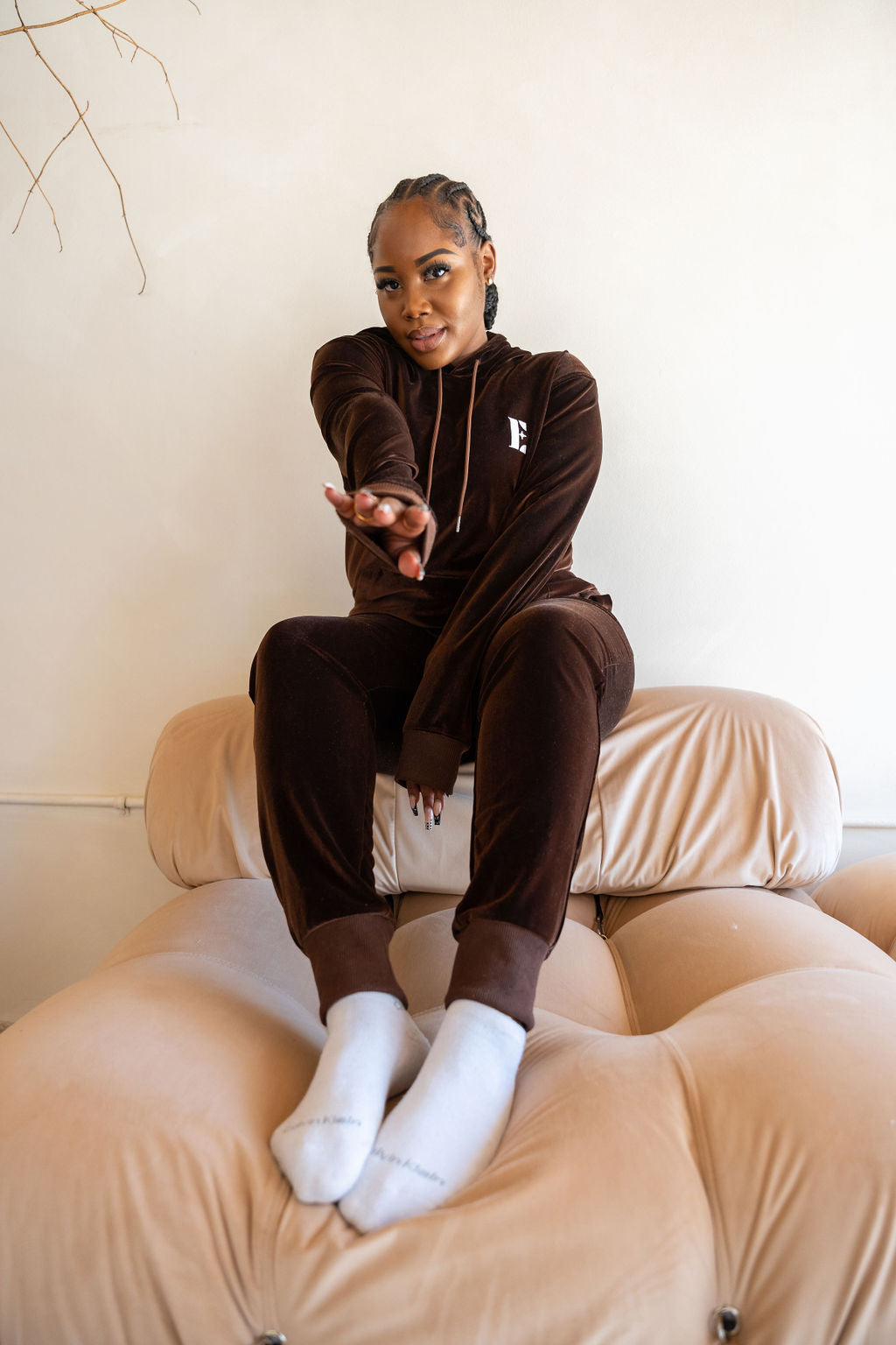 Model sitting on a bed wearing dark brown velvet hoodie and joggers. The hoodie has the E's Element logo imprinted in white. 