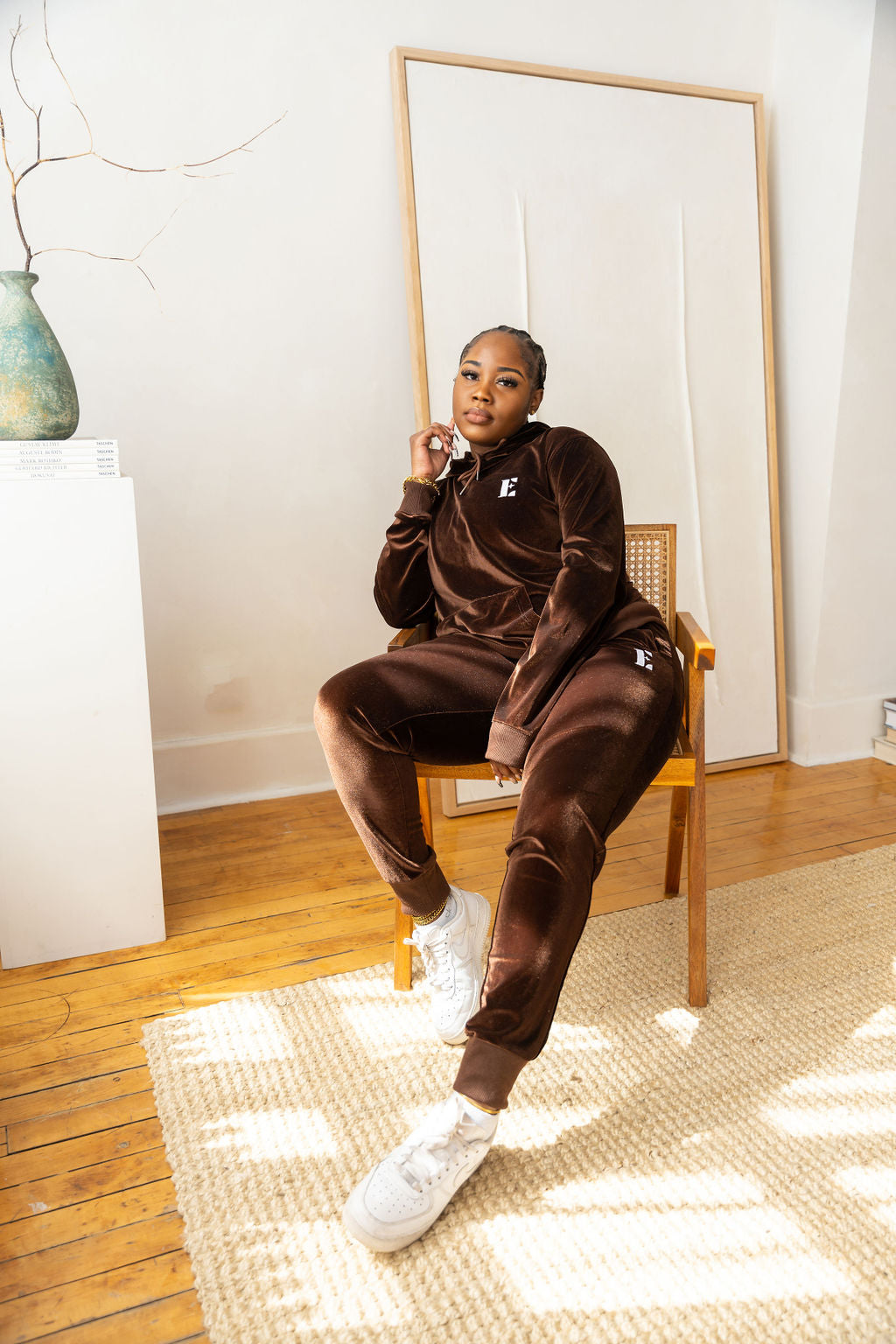 Model sitting on a wooden chair and wearing dark brown velvet hoodie and joggers. The hoodie has the E's Element logo imprinted on it. She is also wearing white sneakers.