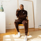 Model sitting on a wooden chair and wearing dark brown velvet hoodie and joggers. The hoodie has the E's Element logo imprinted on it. She is also wearing white sneakers. 
