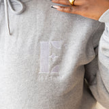 Model wearing a light grey hoodie with the E's Element logo imprinted in white. On her ring and index finger are 18k gold rings. The hoodie is a part of the Essential Light Grey Sweatsuit set by E's Element.