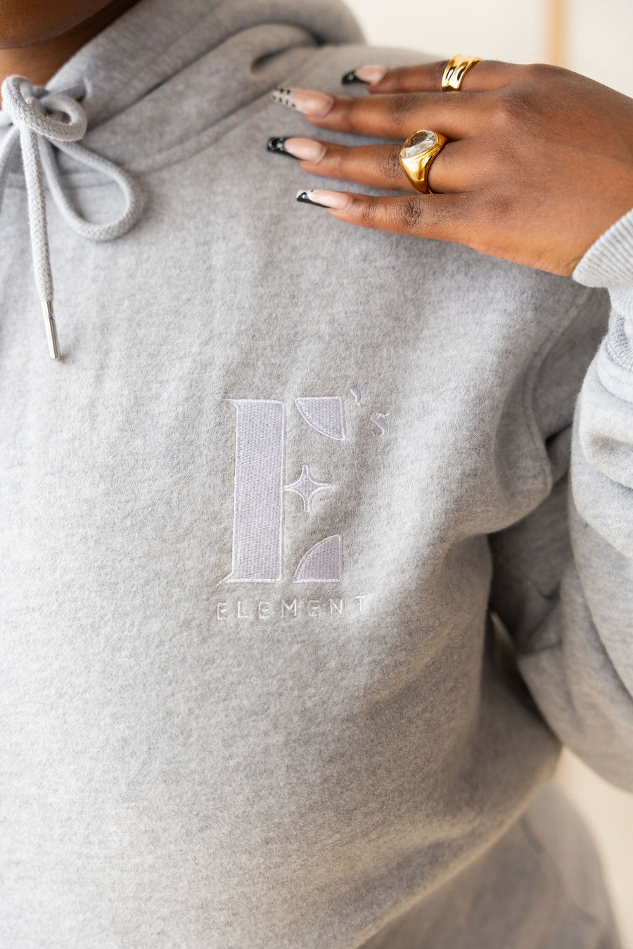 Model wearing a light grey hoodie with the E's Element logo imprinted in white. On her ring and index finger are 18k gold rings. The hoodie is a part of the Essential Light Grey Sweatsuit set by E's Element.