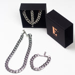 Stainless steel chain bracelet necklace. On the left, the necklace and bracelet are placed in a black opened container. In front of the container is a chain necklace. On the right is the cap for the container with the E's Element logo imprinted in gold. Under the cap is a steel chain bracelet. The Emmanuela Set in Steel by E's Element.