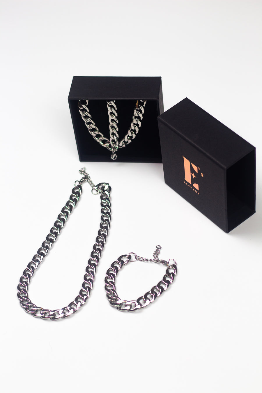 Stainless steel chain bracelet necklace. On the left, the necklace and bracelet are placed in a black opened container. In front of the container is a chain necklace. On the right is the cap for the container with the E's Element logo imprinted in gold. Under the cap is a steel chain bracelet. The Emmanuela Set in Steel by E's Element.