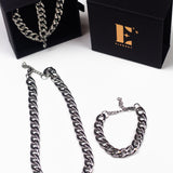 Stainless steel chain bracelet necklace. On the left, the necklace and bracelet are placed in a black opened container. In front of the container is a chain necklace. On the right is the cap for the container with the E's Element logo imprinted in gold. Under the cap is a steel chain bracelet. The Emmanuela Set in Steel by E's Element.