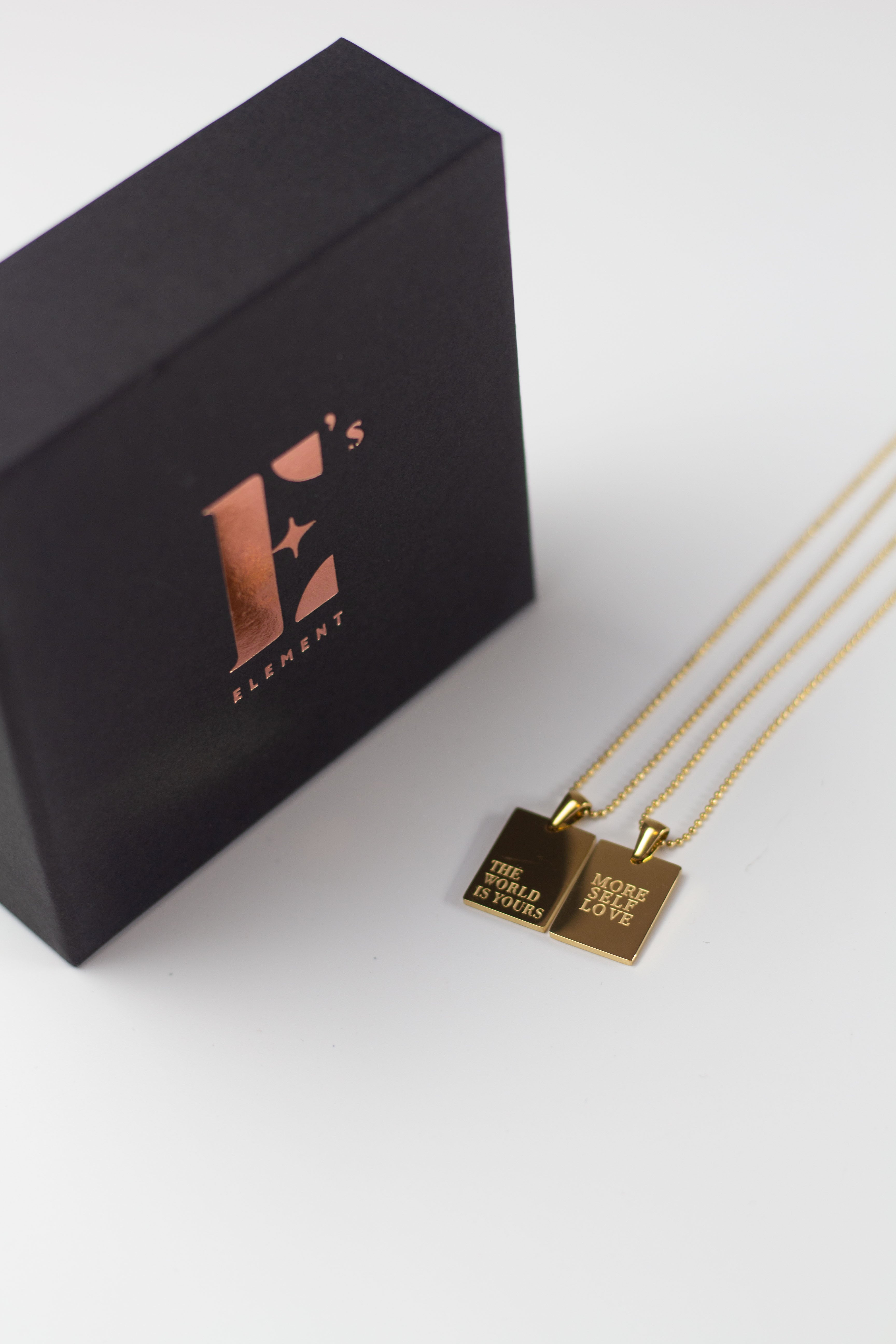 Two 18k gold stainless steel chain necklaces placed side-by-side. One of the necklaces is engraved with the words "THE WORLD IS YOUIRS" and the other with the words "MORE SELF LOVE". E's Element "The World Is Yours" Chain Necklace by E's Element.