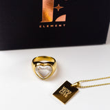18k gold stainless steel ring with a heart-shaped gem in the center. There is an 18k gold stainless steel chain necklace beside it with the words "MORE SELF LOVE" engraved. E's Element "MORE SELF LOVE" Chain Necklace by E's Element.