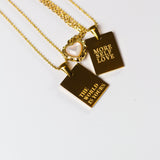 Three 18k gold stainless steel necklaces placed side-by-side. The first and third necklace have the words "THE WORLD IS YOURS" and "MORE SELF LOVE" engraved. The necklace in the middle is heart shaped and has a gem in the center. E's Element "The World Is Yours" Chain Necklace by E's Element.