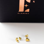 18k gold heart shaped studs. Above it is a black container with E's Element logo imprinted in gold. Ella's Element Dainty Heart Studs by E's Element.
