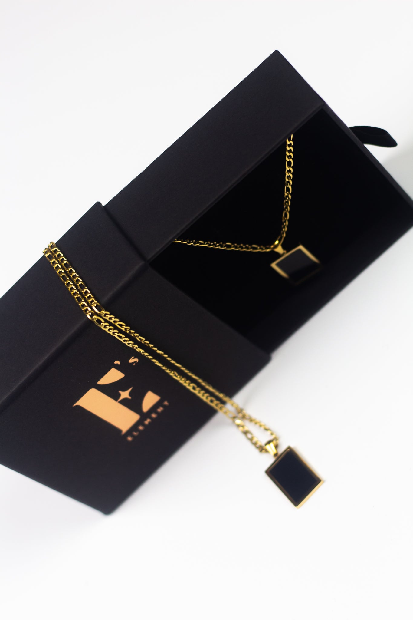 18k gold necklace with a black rectangular charm placed in its container. On the left is the cap for the container with the E's Element imprinted in gold with the necklace on placed on top. The Infinity Zircon Necklace by E's Element.