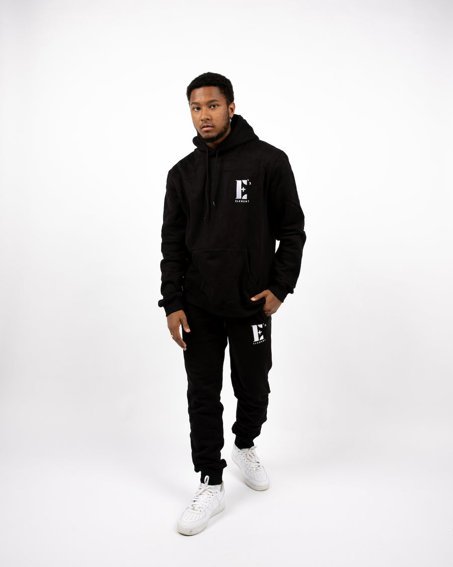 Model wearing a black sweat suit. The hoodie and sweat pants has the E's Element logo imprinted in white. The model is also wearing white sneakers. E's Element Essential Smoky Black Sweatsuit Set by E's Element.