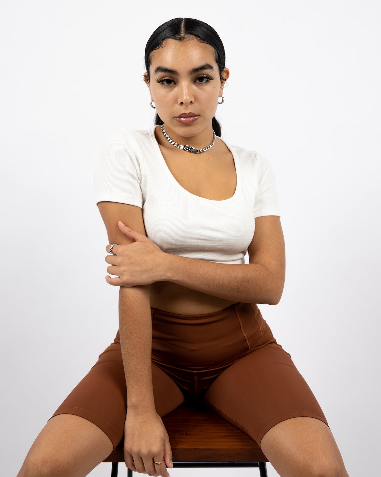 Model sitting on a chair in a white crop top and silver chain necklace and hoop earrings. She is wearing brown biker shorts. The Ella Shaping Cycling Shorts (Petite Size) by E's Element.