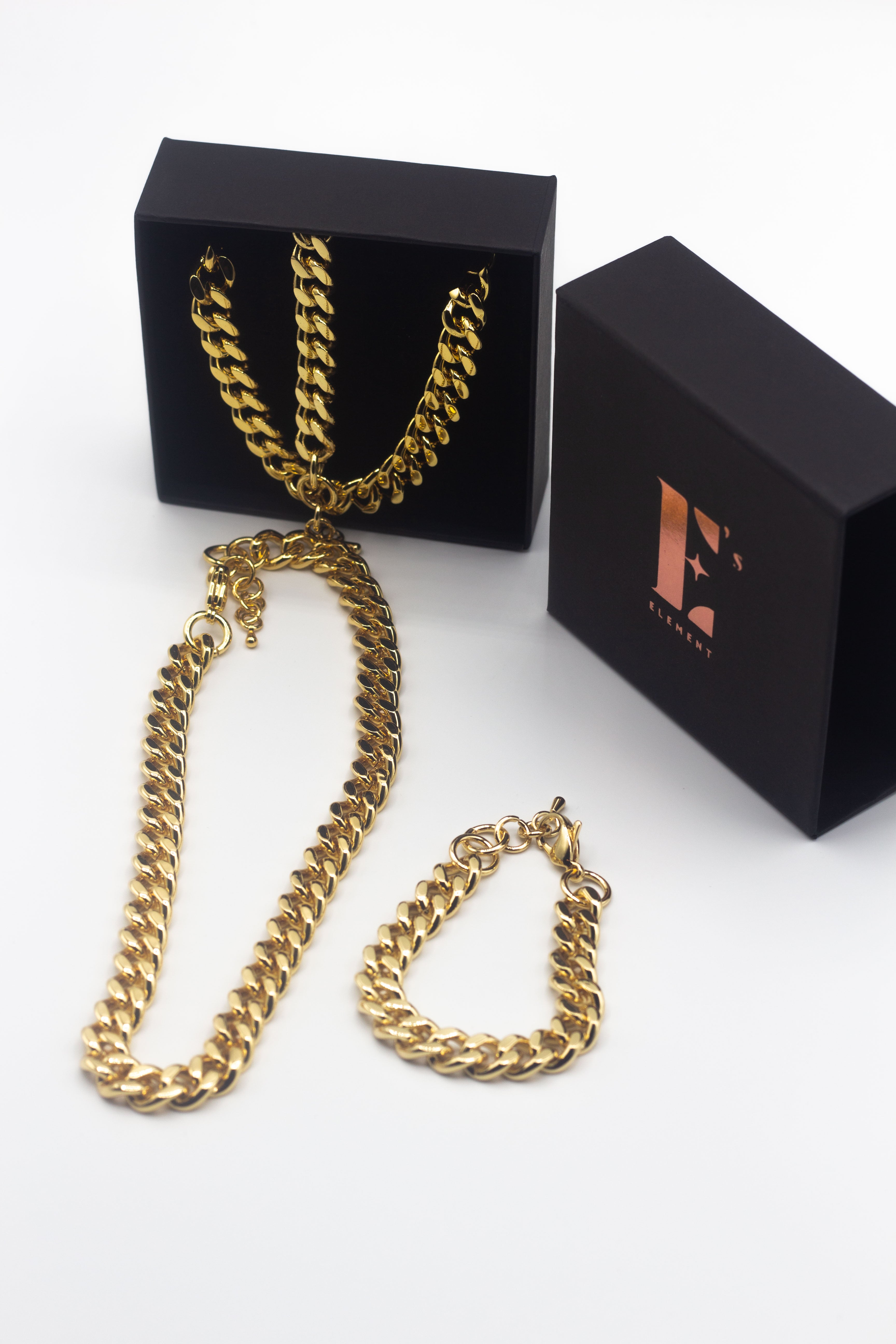 18k gold chain and necklace. On the left, the chain necklace and bracelet are placed in a black opened container. In front of the container is a gold chain necklace. On the right is the cap for the container with the E's Element logo imprinted in gold. Under the cap is a gold chain bracelet. The Emmanuela Set in Gold by E's Element.